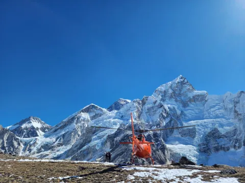 A close-up view of a silver helicopter landed on a snowy plateau at a high-altitude site in the Himalayas with mountain peaks and camping tents in the background.
